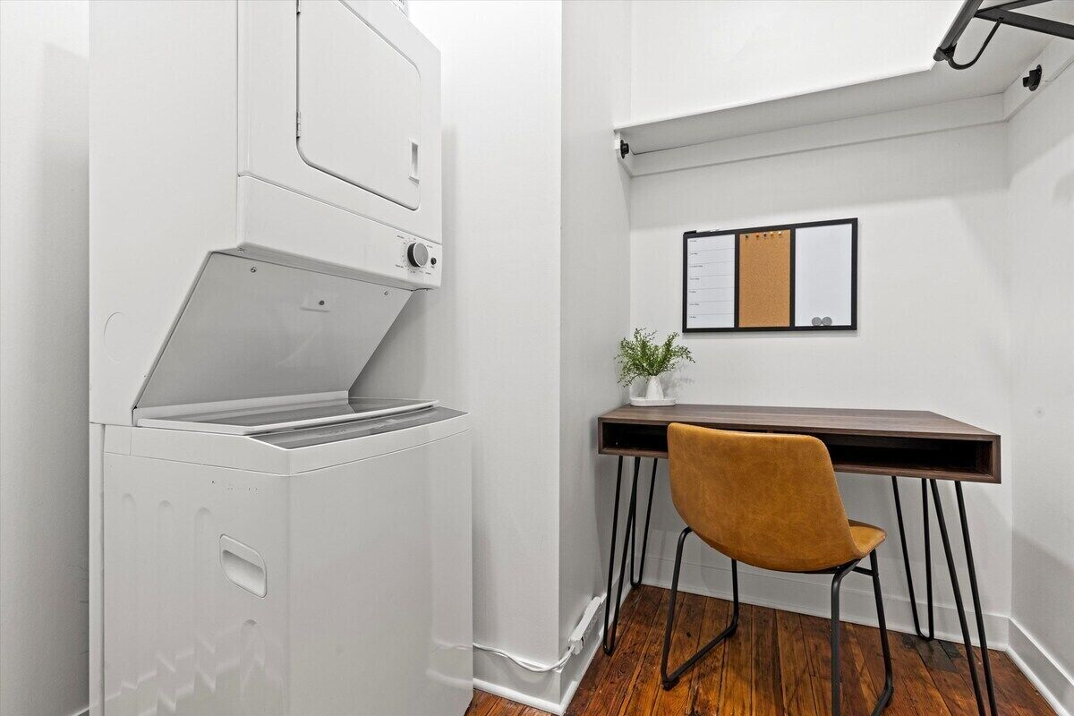 A compact room with a white appliance, a wooden desk, a chair, and a corkboard on the wall.