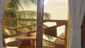 A room with a bed, a wooden bench, and a view of the beach and palm trees through a large window. - AMANI BEACH VILLA (Bwejuu)