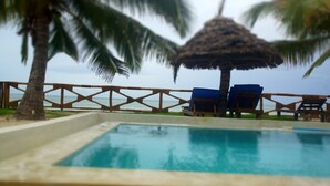 A pool area with a thatched umbrella, two lounge chairs, and a wooden fence. - AMANI BEACH VILLA (Bwejuu)