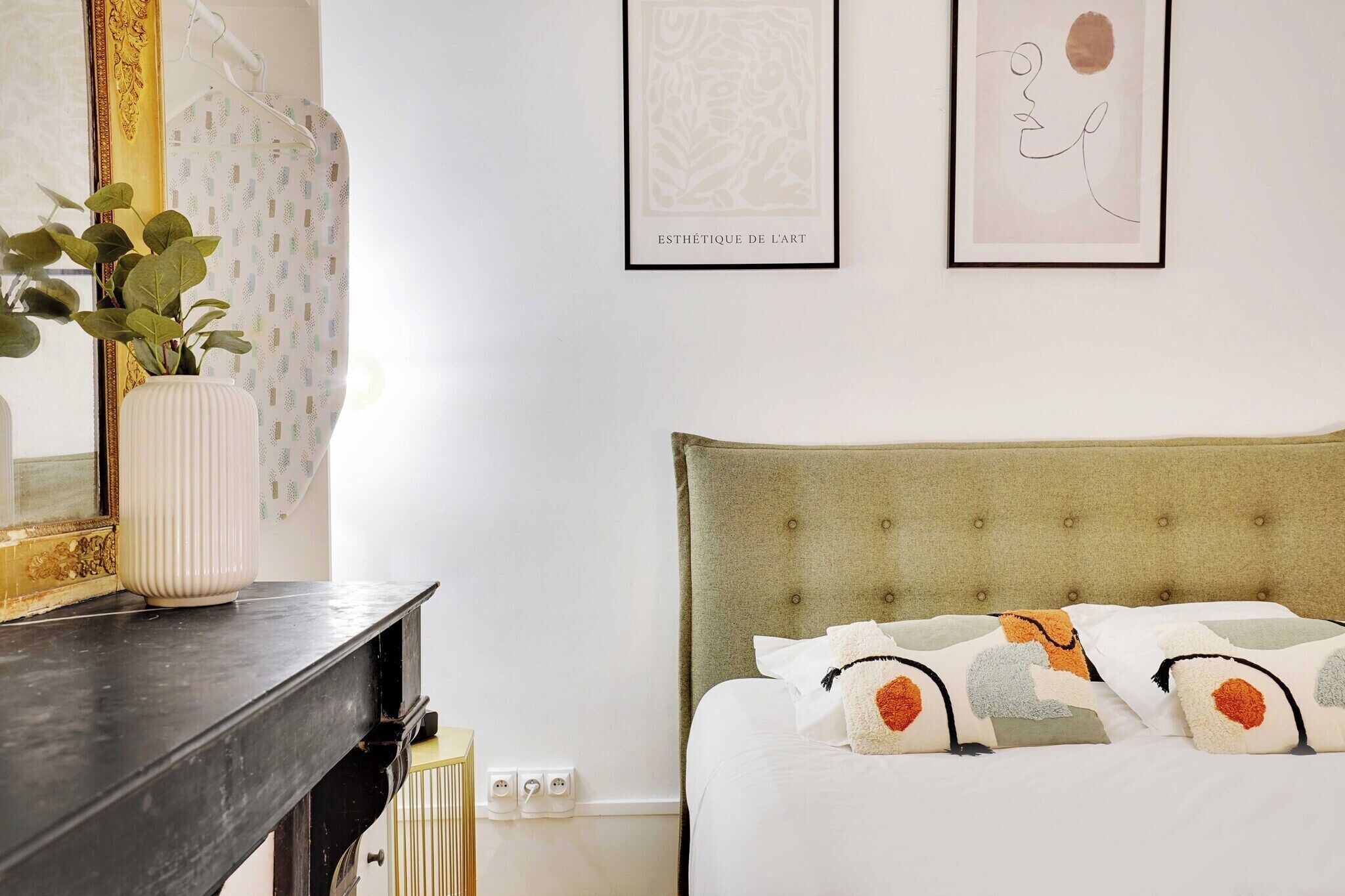A neatly arranged bedroom with a bed, pillows, a bedside table, a mirror, and wall art.