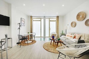 A modern living room with a sofa, a dining table, and chairs, a TV mounted on the wall, and large windows with curtains. - Charming apartment - 1BR/5P - Opéra Garnier (Paris)