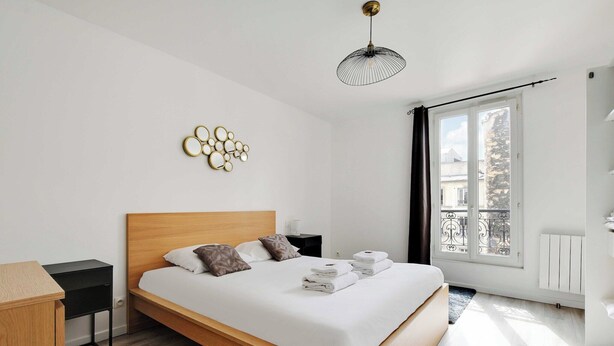 Quarto de hotel moderno com cama de madeira, uma secretária pequena, um objeto decorativo na parede e uma janela grande com vista para edifícios.