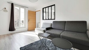 Uma sala de estar moderna com um sofá cinzento, uma mesa pequena e uma cadeira. Há uma janela com cortinas, um radiador e um candeeiro de teto.