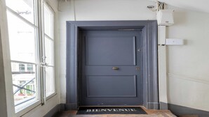 Una puerta gris oscuro con un felpudo que dice "BIENVENUE" en la entrada, una ventana a la izquierda y un equipo de aire acondicionado en la pared.