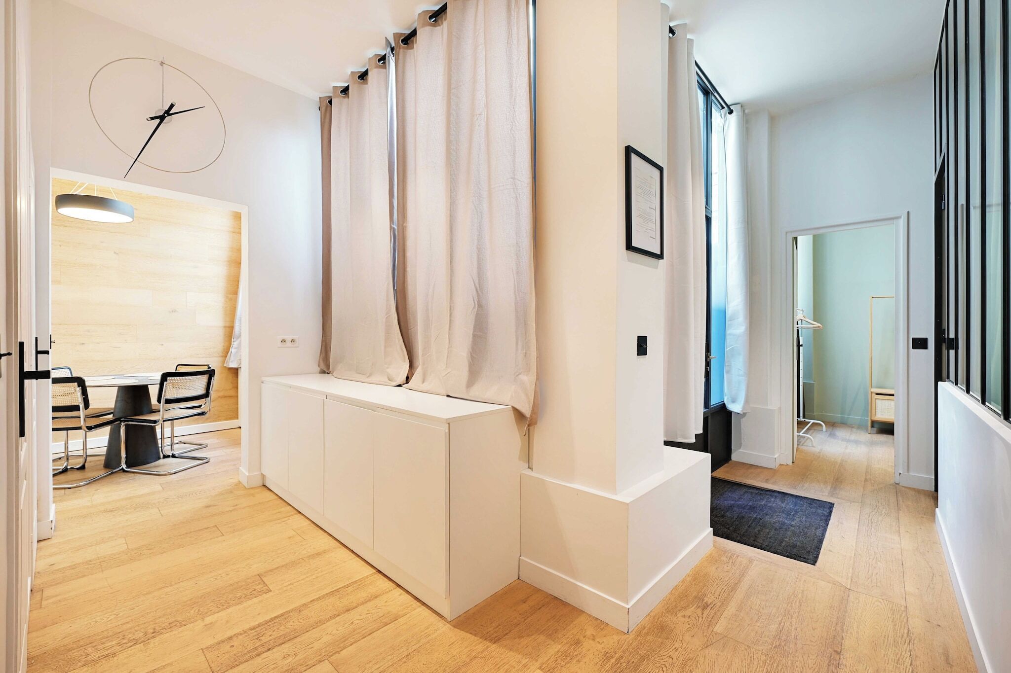 A modern hotel room with a wooden floor, a white console table, a black chair, a round wall clock, and a large window with curtains.