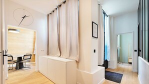 A modern hotel room with a wooden floor, a white console table, a black chair, a round wall clock, and a large window with curtains. - Haven of peace near the Palais de Garnier (Paris)