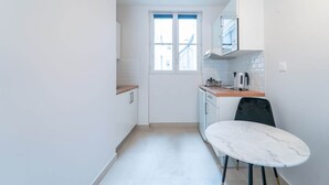 A compact kitchen with white cabinets, a window, and a small dining area with a white table and two chairs. - Charming studio- 2P- Jardin des plantes/Austerlitz (Paris)
