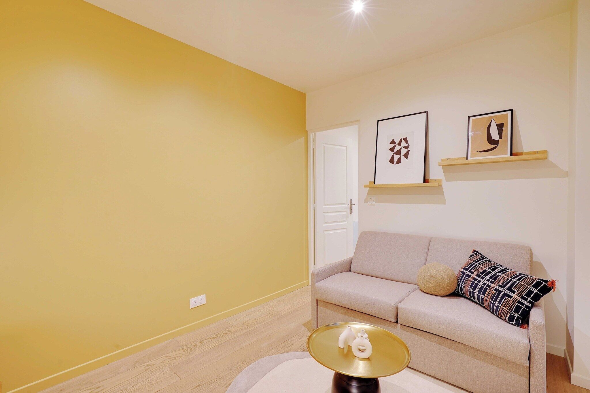 A living room with a beige sofa, a round coffee table, and two framed artworks on a shelf above the sofa.