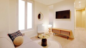 Un salon moderne comprenant un canapé, une table basse ronde, une table d’appoint avec une lampe, un téléviseur fixé au mur et une fenêtre avec des stores.
