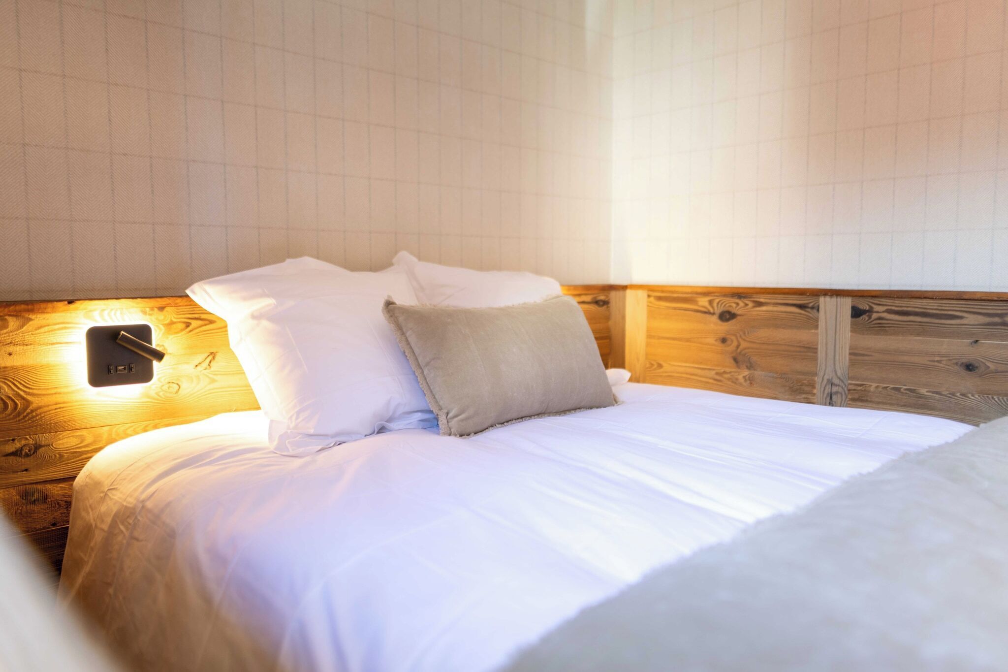 A neatly made bed with white linens and two pillows, a wooden headboard, and a wall-mounted light fixture.