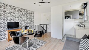Ein modernes Wohnzimmer mit Flachbildfernseher, schwarz-weiß gemustertem Wandpapier, einem Essbereich mit Tisch und Stühlen sowie einer Küche mit Mikrowelle und Spüle.