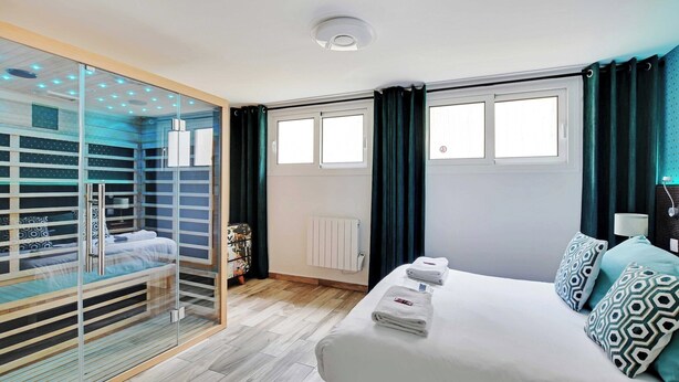 Ein modernes Hotelzimmer mit einem gläsernen begehbaren Kleiderschrank, einem großen Bett mit gemusterten Kissen, einem Nachttisch mit Lampe und einem Vorhang vor dem Fenster.