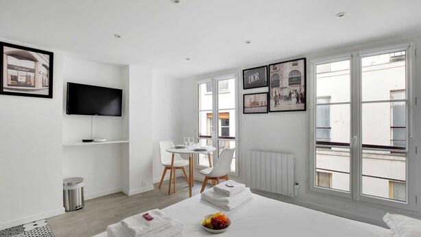 A modern hotel room with a bed, a TV, a dining table with chairs, and large windows.