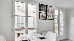 A dining area with a round table set for two, white chairs, and a window with a view of buildings.