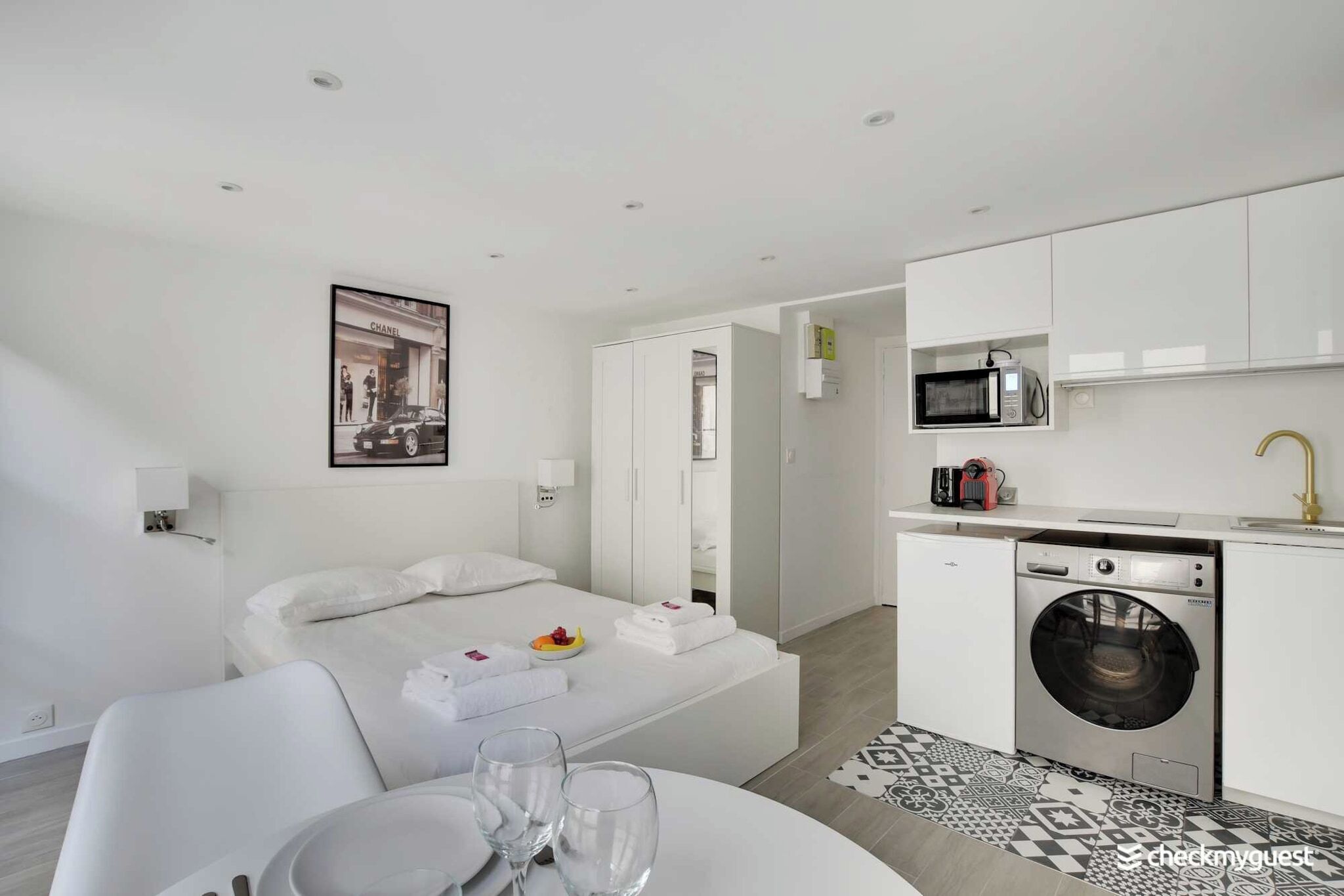 Et moderne hotelværelse med seng, spiseplads med bord og stole, et køkken med vaskemaskine og et badeværelse, der kan ses gennem en åben dør.