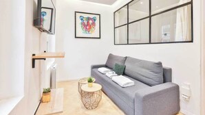 A modern living room with a grey sofa, a mounted TV, a framed artwork, and a large window. - Classy apartment in the heart of Le Marais (Paris)