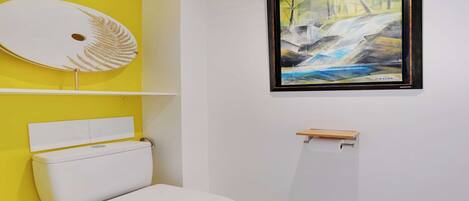 Une salle de bain avec un mur d’accent jaune, des toilettes blanches, un tableau encadré et un lavabo blanc avec une étagère en bois.