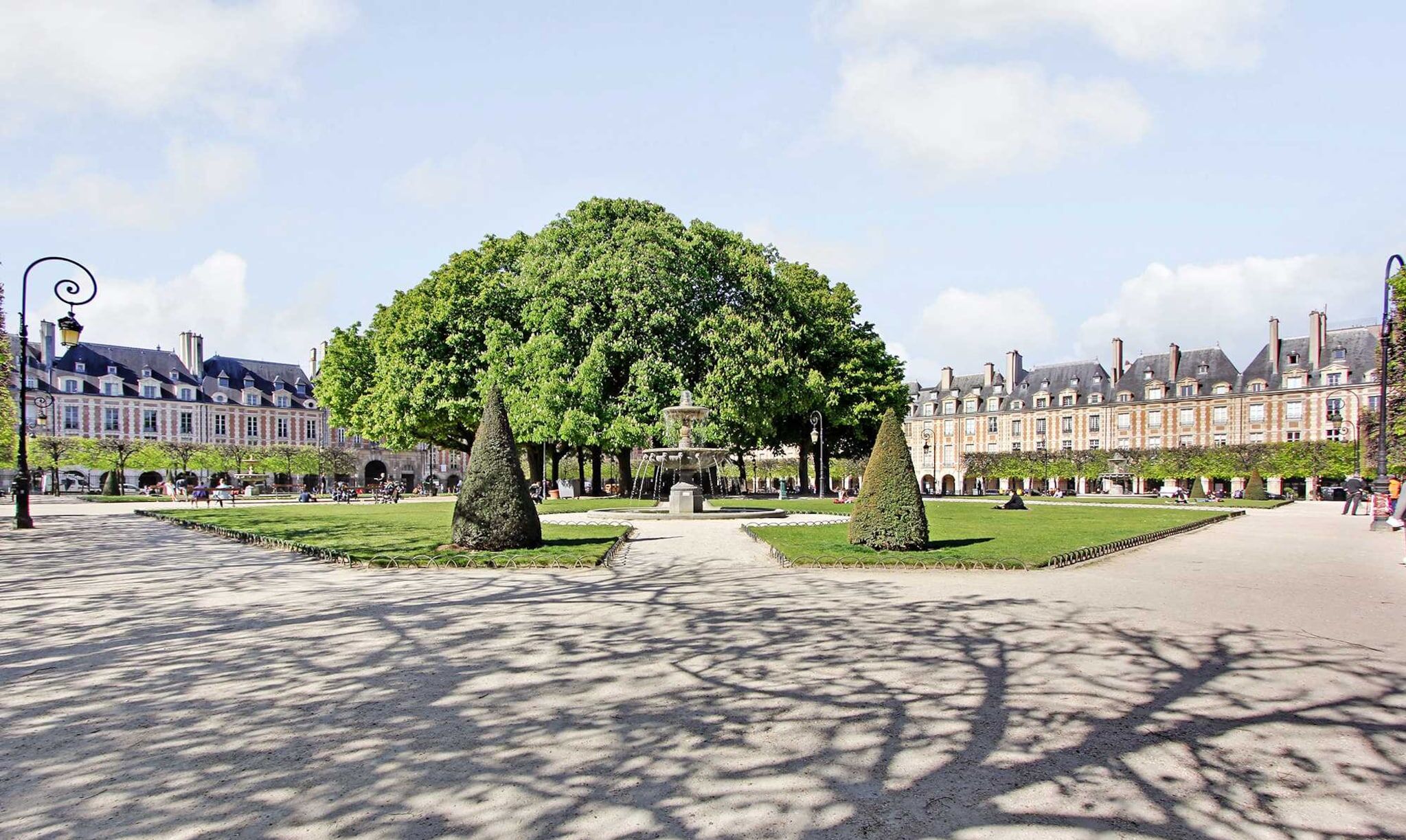 Une vaste place agrémentée d’une fontaine centrale, entourée de pelouses et d’arbres aux formes soignées.