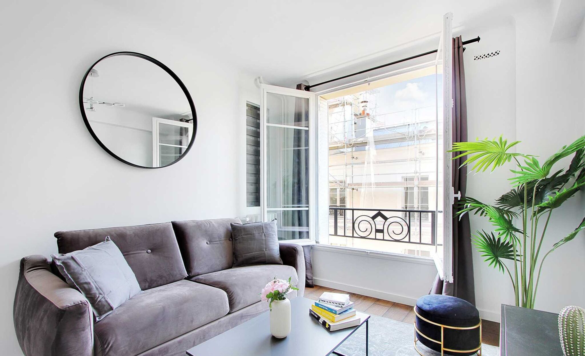 A modern living room with a grey sofa, a round mirror, a coffee table with books, a potted plant, and a balcony with a railing.