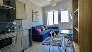 Un espacio habitable compacto con kitchenette, un sofá azul con cojines de colores, una pequeña mesa de comedor y un ventanal que proporciona luz natural.