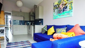 Una sala moderna con un sofá azul, almohadones coloridos, un tapete estampado y una cocina al fondo.