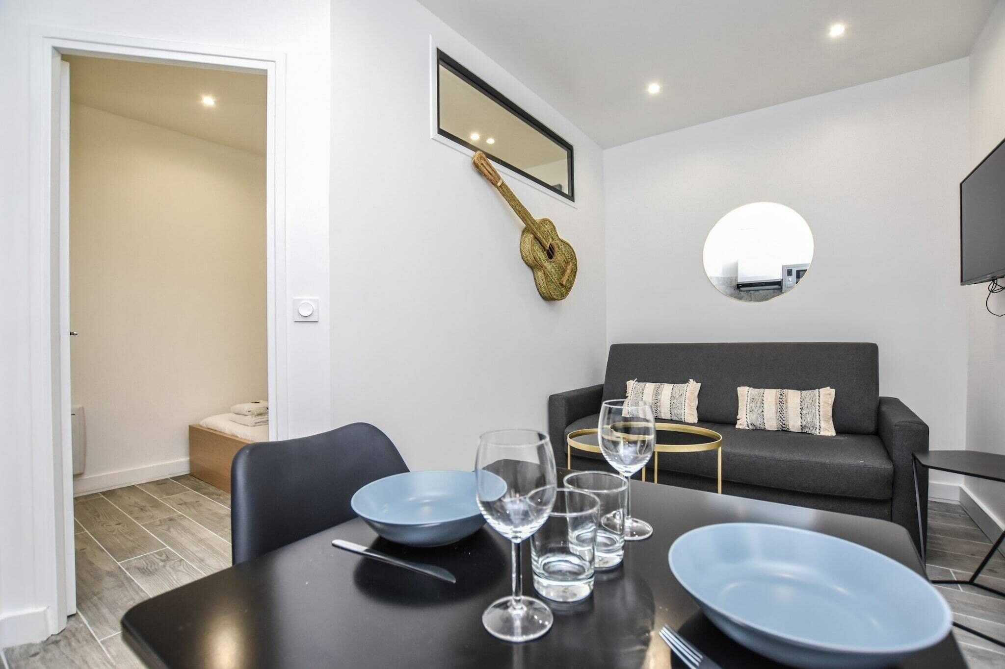 Un salon moderne avec une table à manger dressée pour quatre personnes, un canapé gris, un petit miroir rond et une guitare accrochée au mur.