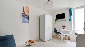 A modern, well-lit room with a white wardrobe, a dining table with two chairs, a small round table with a cup and pastries, and a large window with curtains. - Cozy Studio in the heart of the Marais (Paris)