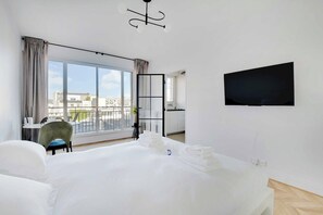 Et hotelværelse med en stor seng, et skrivebord med stol, et fjernsyn ophængt på væggen og en balkon med udsigt over byen.