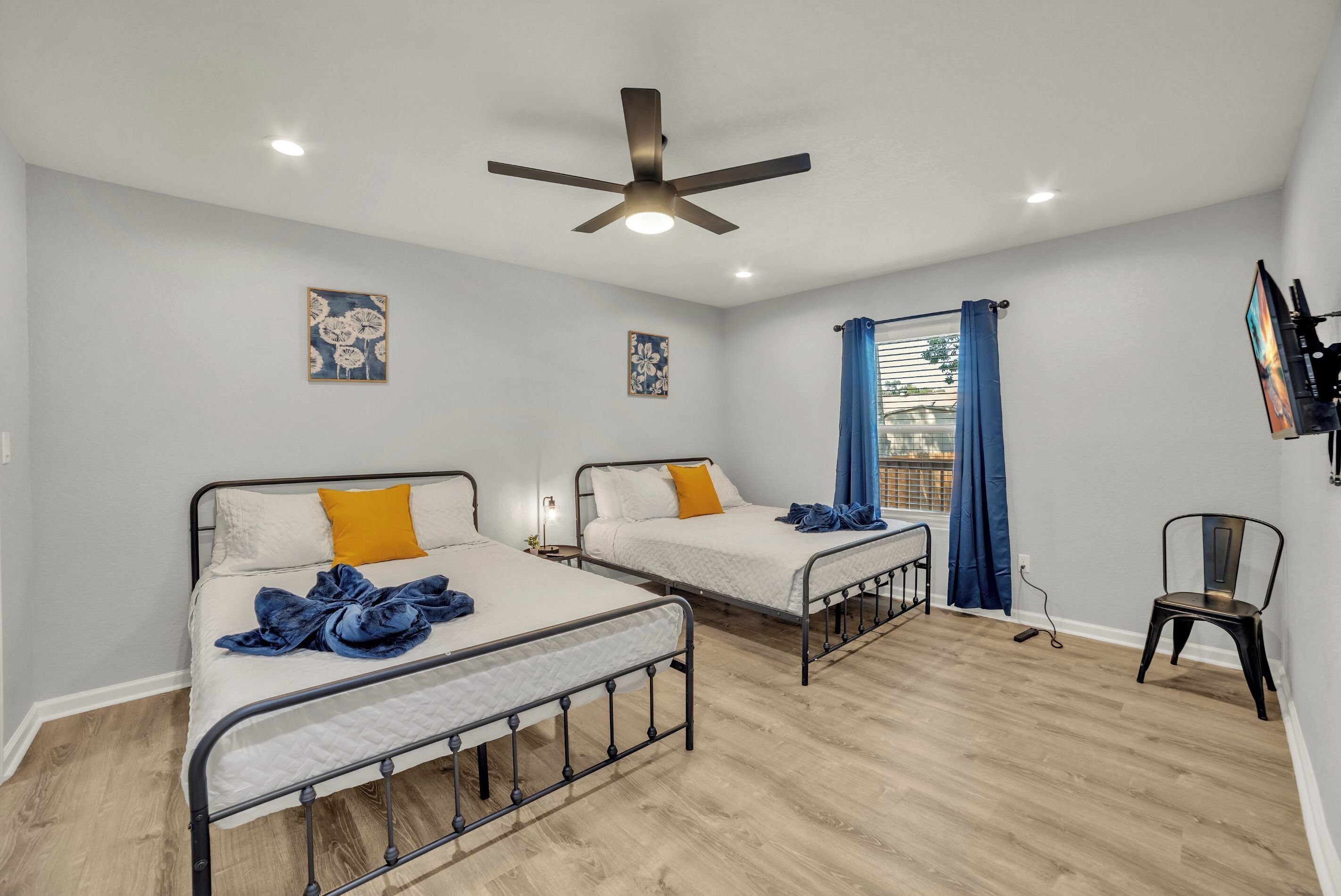 A room with two beds, a ceiling fan, artwork on the walls, a window with curtains, and a chair.