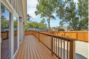 Una terraza de madera con barandilla, ventanas grandes y vistas a árboles y una valla.