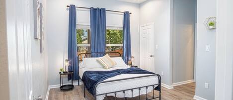 Une chambre à coucher avec un lit, des rideaux, un ventilateur de plafond, une table de chevet et une fenêtre donnant sur un paysage verdoyant.