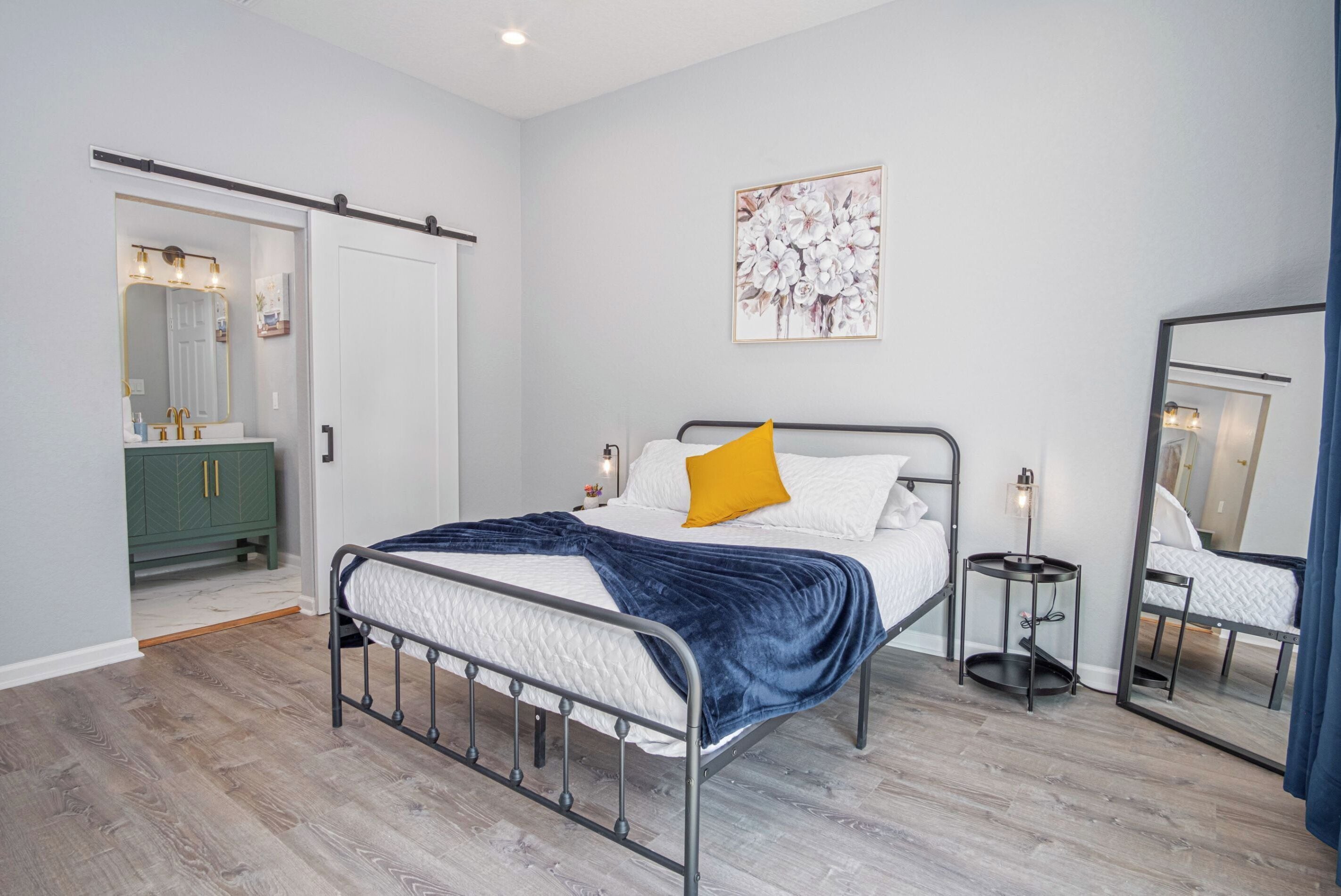 A modern bedroom with a metal bed frame, a white bedspread, a blue blanket, and a yellow pillow. There is a green dresser with a mirror, a small bedside table with a lamp, and a wall-mounted artwork.