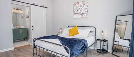 A modern bedroom with a metal bed frame, a white bedspread, a blue blanket, and a yellow pillow. There is a green dresser with a mirror, a small bedside table with a lamp, and a wall-mounted artwork.