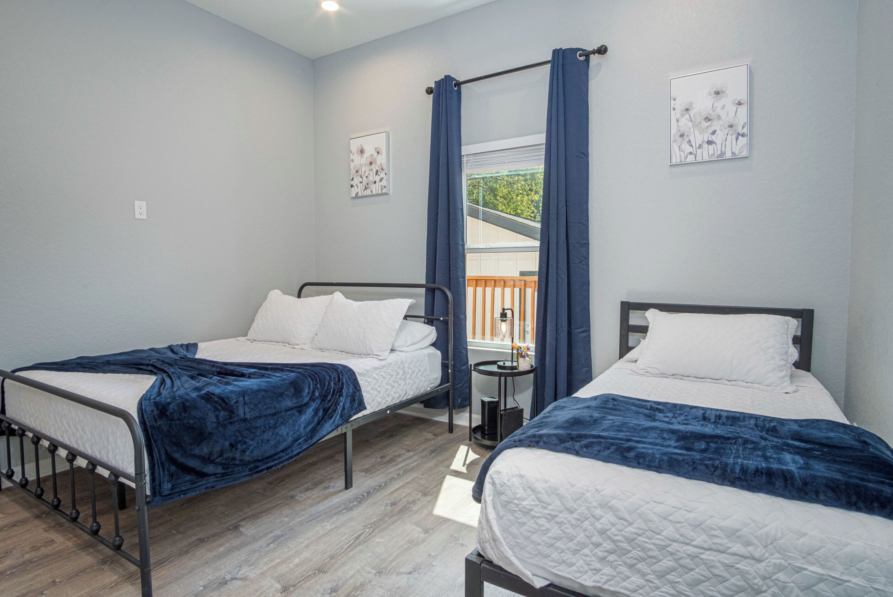 A bedroom with two beds, a window with curtains, and a small table with a lamp.