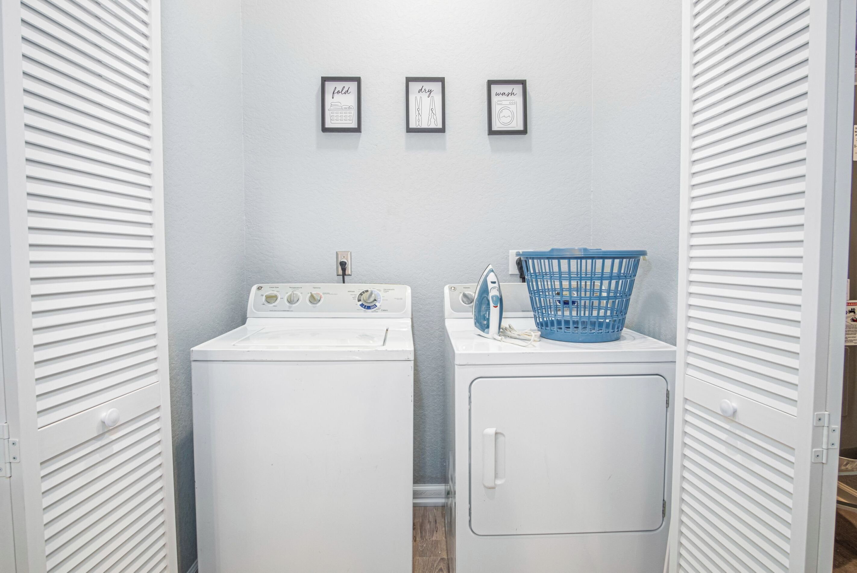 A laundry room with a front-loading washer and dryer, a blue laundry basket, and three framed pictures on the wall.