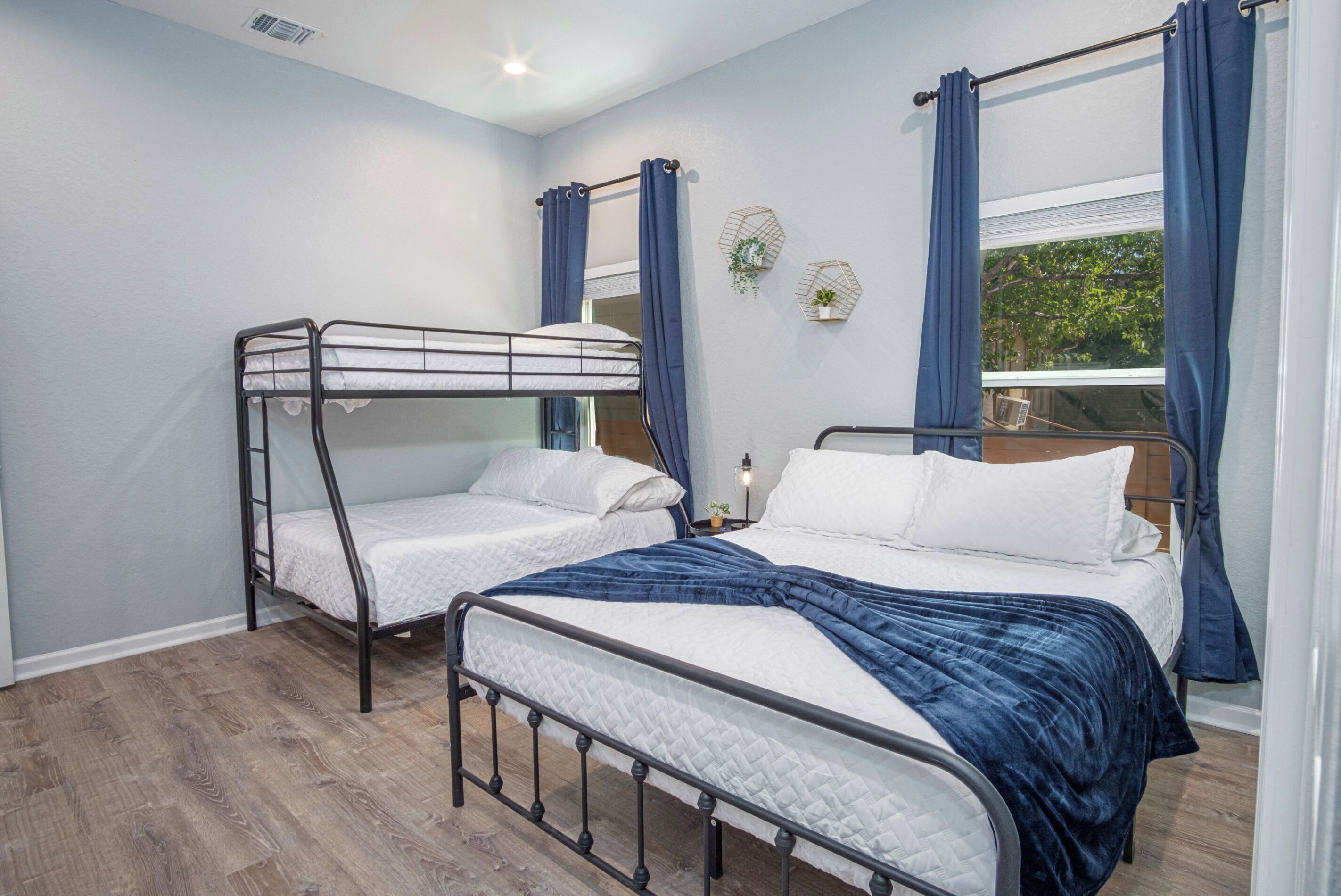 Een kamer met twee stapelbedden, een raam met gordijnen, een klein tafeltje met een lamp en een wandplank met planten.