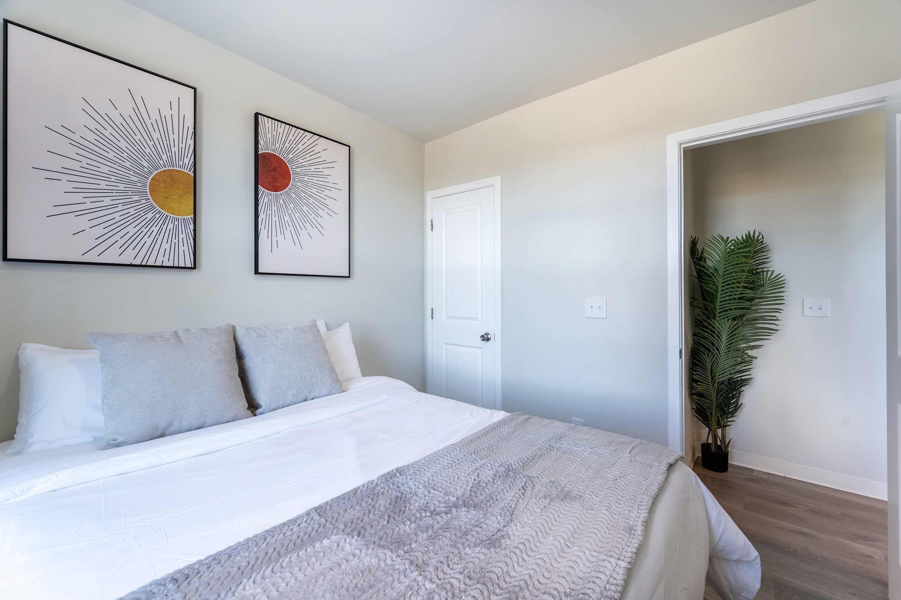 Une chambre à coucher avec un lit, deux œuvres d’art accrochées au mur, une porte et une plante en pot.