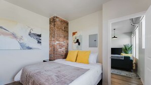 Une chambre dotée d’un grand lit, d’un mur décoratif en briques et donnant sur un espace de vie comprenant un canapé et des plantes.