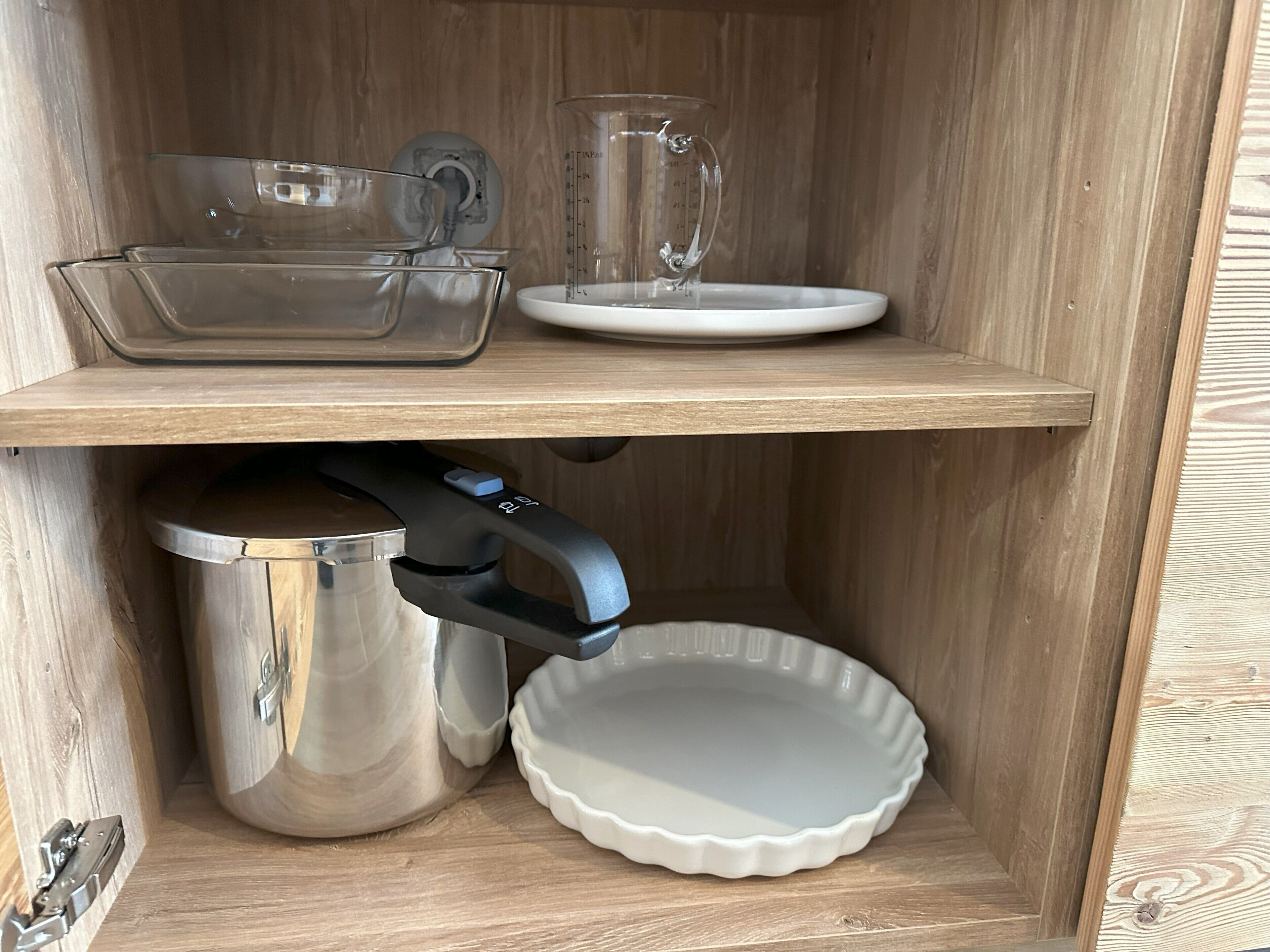 A wooden cabinet with two shelves, one holding a glass measuring cup, a glass, and a white plate, and the other containing a stainless steel pot, a white plate, and a black appliance.