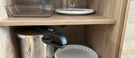 A wooden cabinet with two shelves, one holding a glass measuring cup, a glass, and a white plate, and the other containing a stainless steel pot, a white plate, and a black appliance.