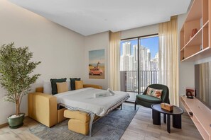 Et moderne hotelværelse med seng, sofa, lænestol, en potteplante og udsigt over bybilledet.