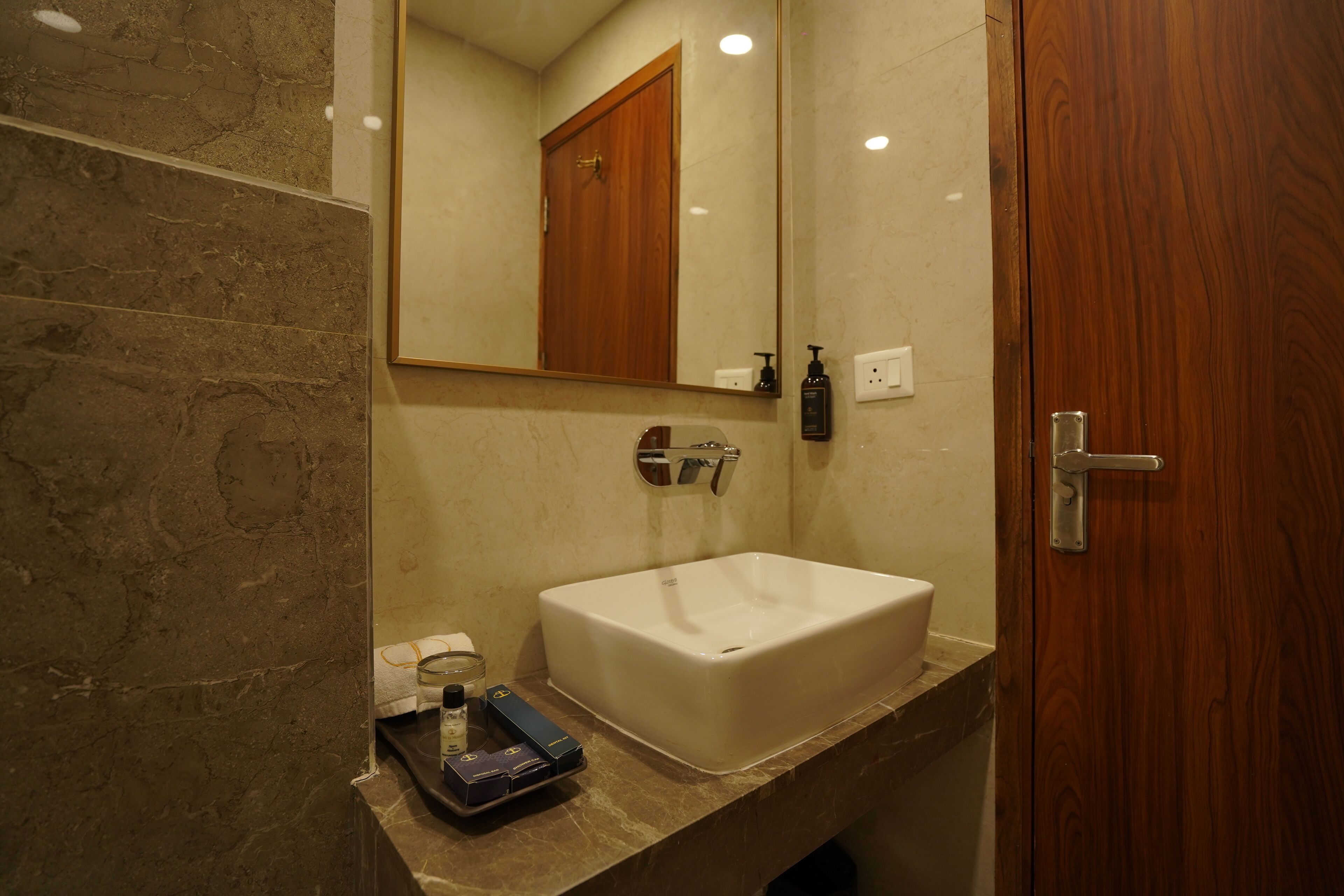 Une salle de bain avec un comptoir en marbre, un lavabo blanc, un miroir, une porte en bois et un flacon de savon pour les mains.