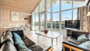 Een ruime woonkamer met leren banken, een centrale salontafel en een televisie. Grote ramen bieden uitzicht op de omgeving.