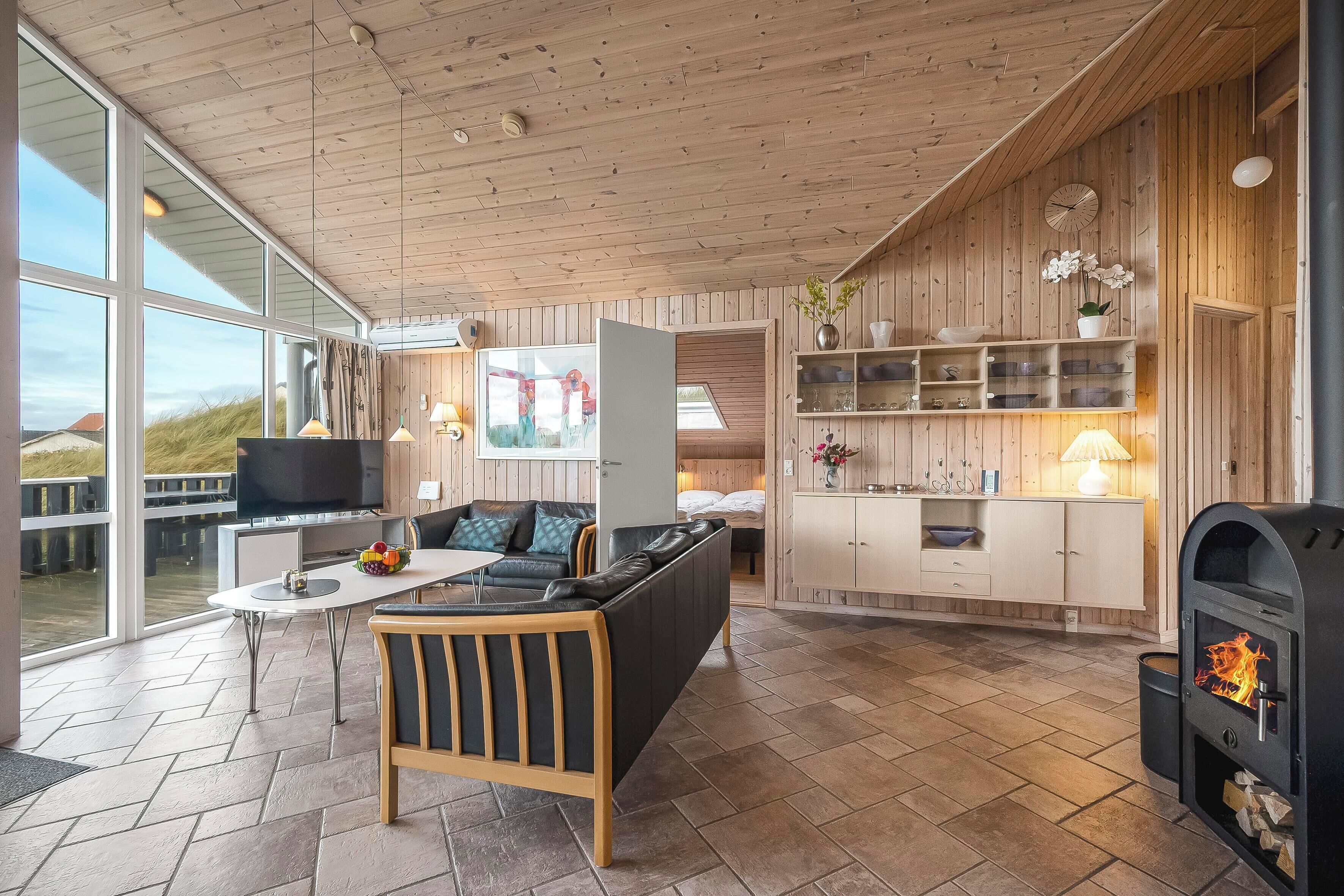 Un salon moderne avec un canapé, une table basse et un téléviseur. Il est doté de grandes fenêtres, de murs en bois et d’une cheminée.