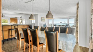 A modern dining area with a large table, multiple chairs, pendant lighting, and a view of the sea.