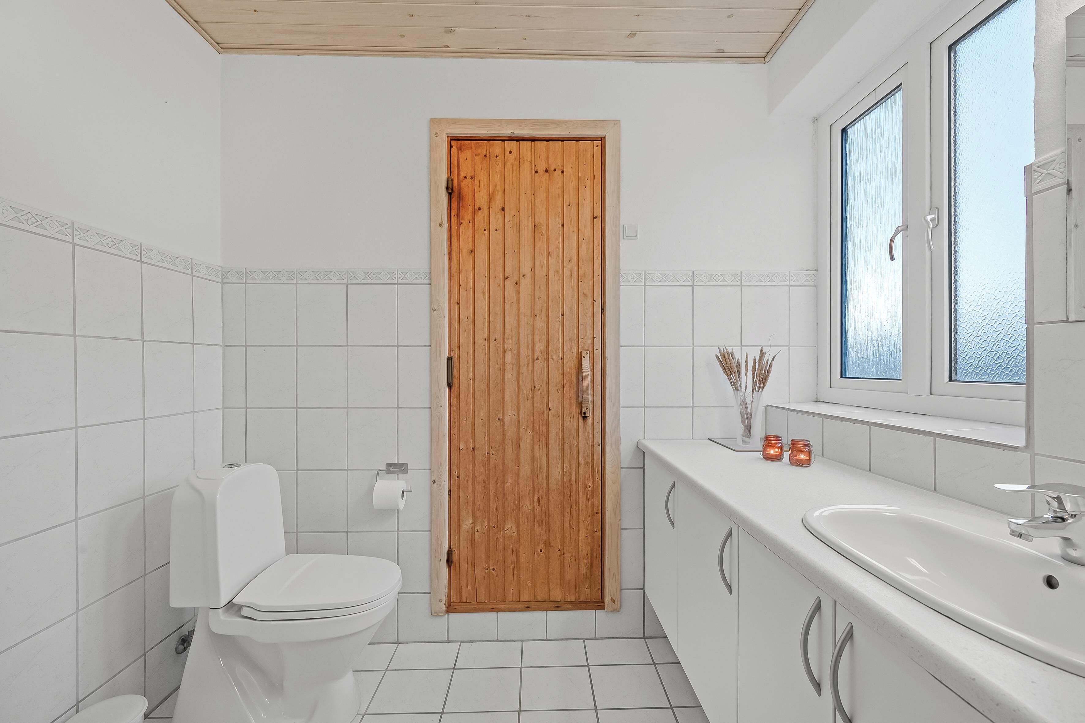 Une salle de bain avec une porte en bois, des carreaux blancs, des toilettes, un lavabo et une fenêtre.