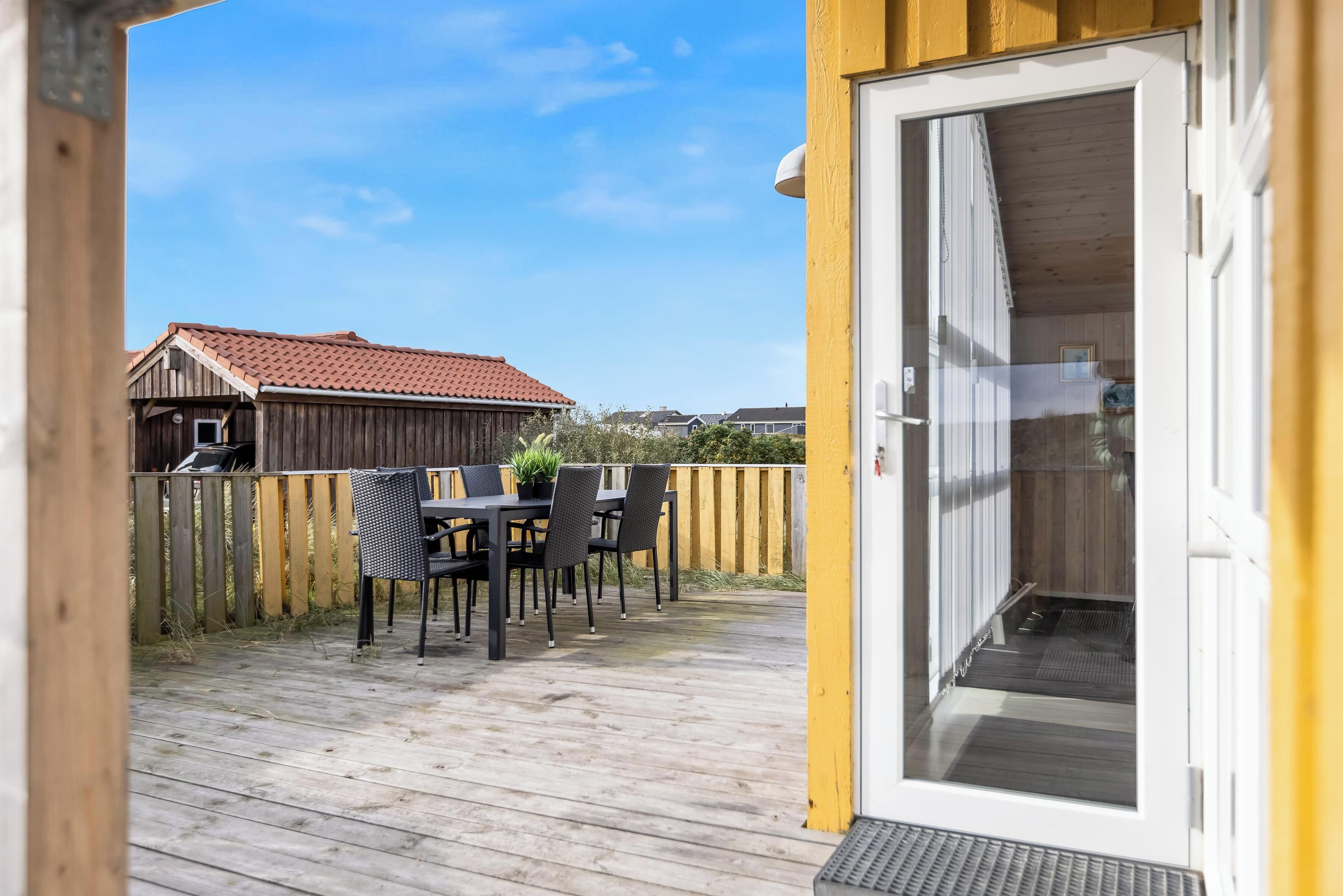 Eine Holzterrasse mit Tisch und Stühlen, eine Glasschiebetür und im Hintergrund ein Gebäude mit rotem Dach.