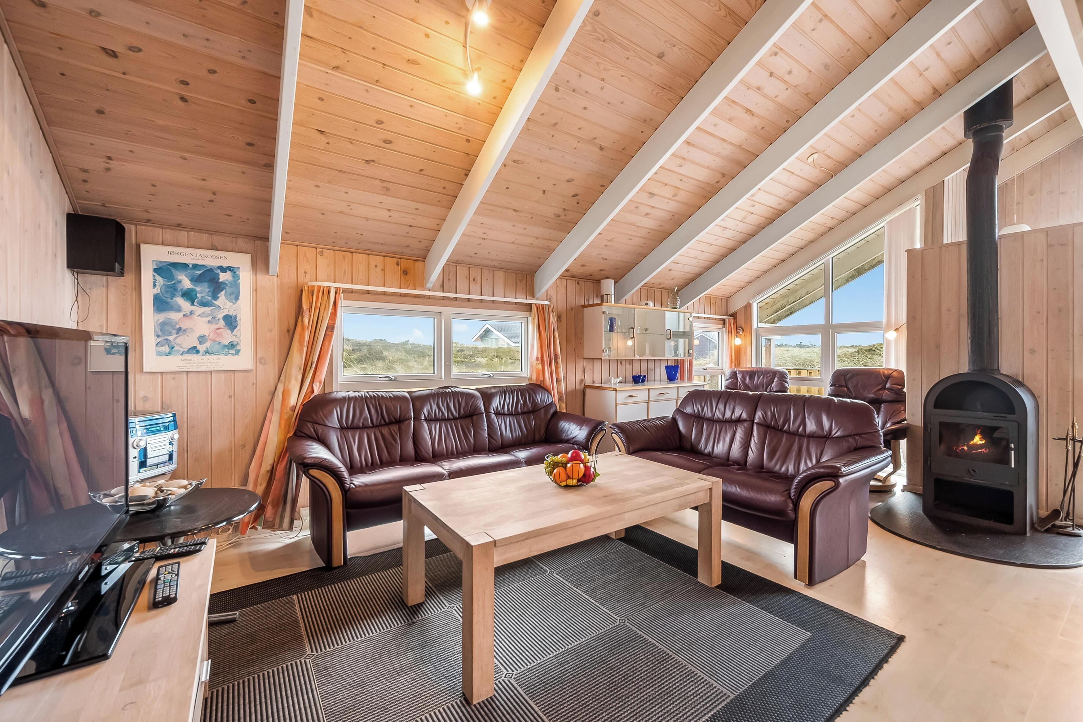 Ein gemütliches Cabin-Interieur mit Holzbalkendecke, Ledersofas, einem zentralen Tisch mit Obst, einem Fernseher und einem Kamin.