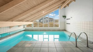 En indendørs swimmingpool med skrå træloft, stort vinduesparti med frit udsyn og flisegulv.