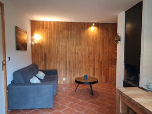 Una habitación con una pared de madera, un sofá gris, una mesa redonda con un objeto azul y una chimenea.